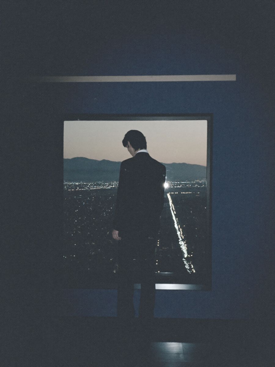 A figure at a high window at dusk, looking out over a city.
